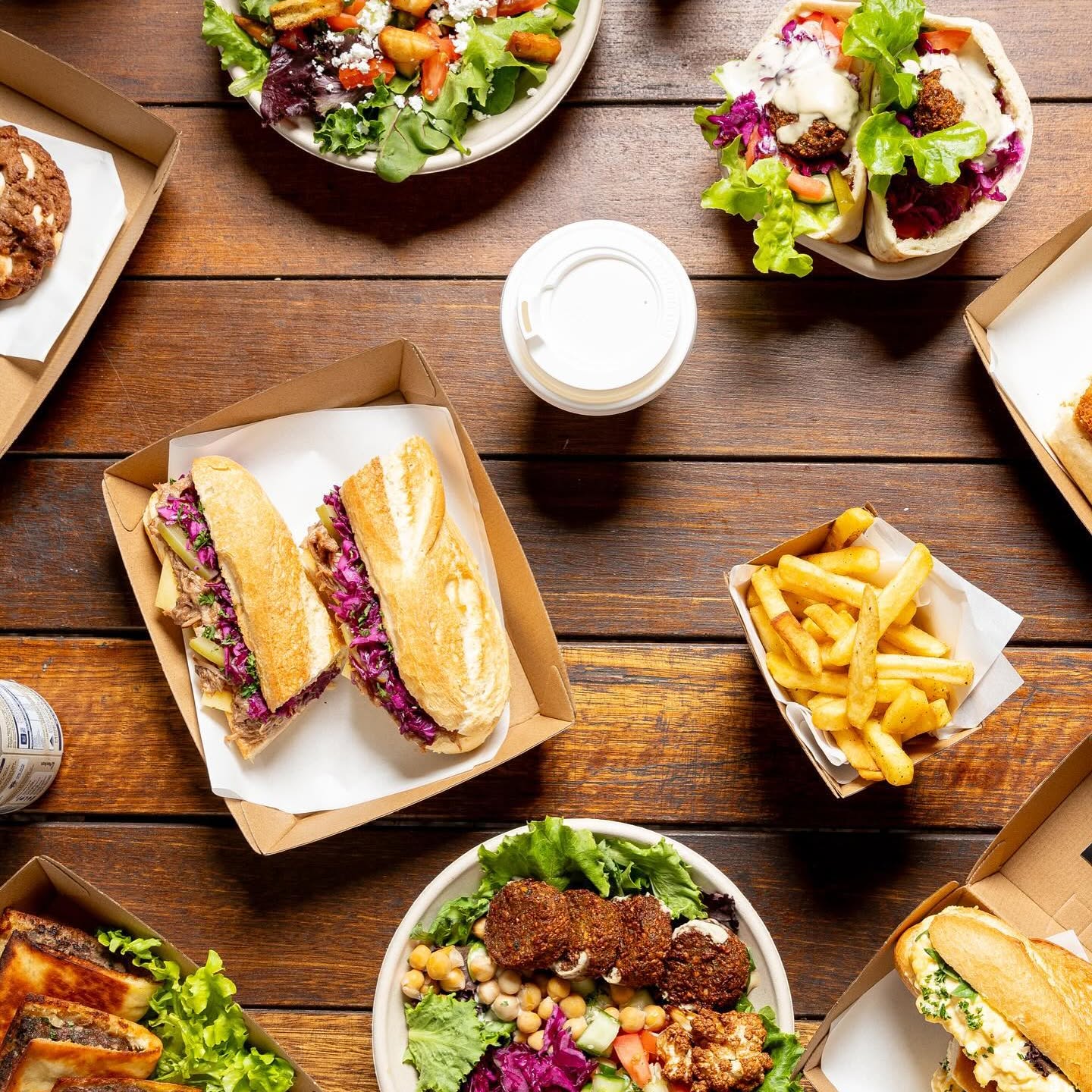 Overhead spread of Fala Fels baguettes, falafel bowl, chips, cookies and pockets on a timber table